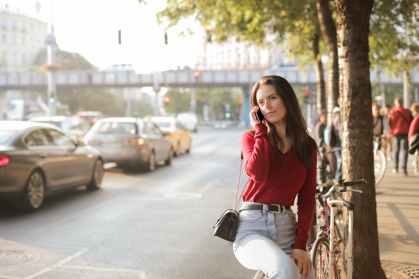 selective focus photo of woman in red long sleeve sweater and blue jeans sitting on metal railing while talking on the phone